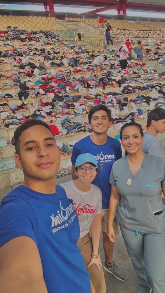 Cuatro misioneros en las graderías de un estadio convertido en centro de acopio. Detrás de ellos, las gradas cubiertas de ropa donada, clasificada en montones que suben hasta arriba, y otras personas organizándola.